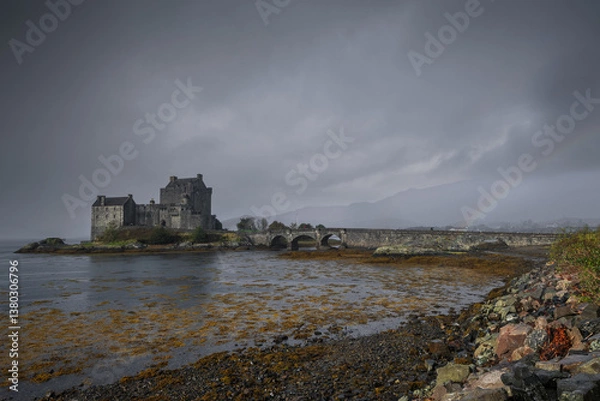 Fototapeta Eilean Donan Castle stands majestically amid autumn hues, its reflection shimmering in calm waters. Misty Highlands and golden trees create a scene of timeless beauty.