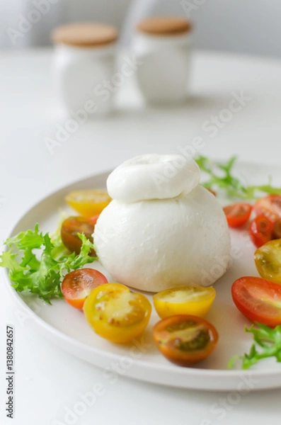 Fototapeta Buratta on a gray plate with cherry tomatoes and herbs on a white table. Traditional Italian food concept. Vertical orientation. Selective focus.