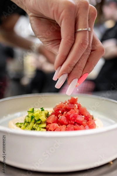 Obraz A hand with manicured nails elegantly sprinkles sesame seeds over a bowl of diced tomatoes and cucumbers, enhancing the fresh, colorful dish. The image captures culinary artistry and food preparation.