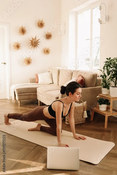Fototapeta A fitness woman does sports at home using a mobile phone and a tripod while sitting on a exercise mat in a cozy interior. The influencer records an online workout. Healthy lifestyle. Selective focus