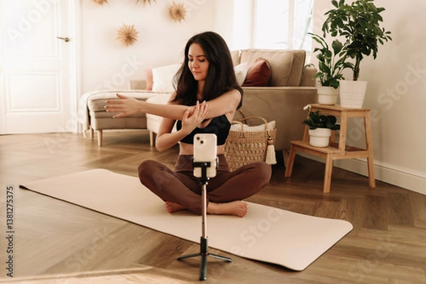 Fototapeta A fitness woman does sports at home using a mobile phone and a tripod while sitting on a exercise mat in a cozy interior. The influencer records an online workout. Healthy lifestyle. Selective focus