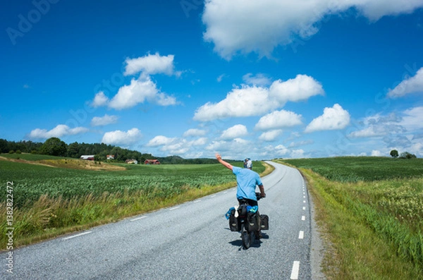 Fototapeta A touring cyclist rides along a scenic countryside road in Västra Götaland County, Sweden, waving joyfully. Open landscape with green fields and bright blue sky creates perfect summer adventure scene