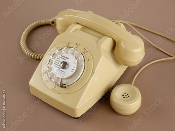 Obraz Un téléphone à cadran rotatif de couleur beige, avec écouteur secondaire, avec ses câbles. Des chiffres et des lettres pour la numérotation sont visibles. Sur fond marron.