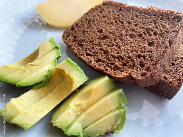 Fototapeta Two square toasts of black bread, sliced ​​green ripe avocado and pieces of hard cheese lie on a round gray textured plate, close-up, top view. Proper nutrition. Vitamins. Healthy snack.