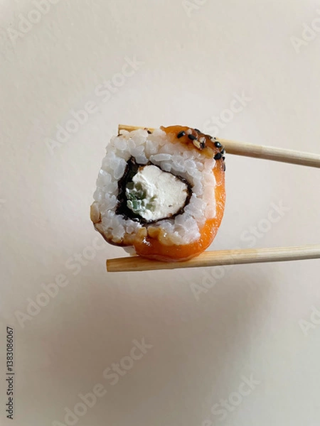 Fototapeta Small square piece of roll with rice, cheese, cucumber, nori and salmon is clamped between two wooden light brown chopsticks on beige background, close-up, side view. Japanese cuisine.