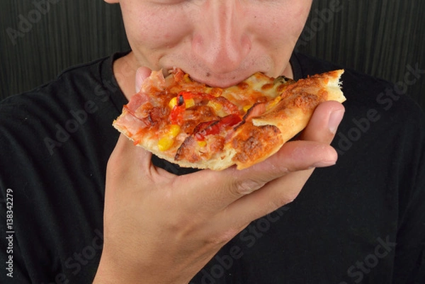 Obraz Portrait of a greedy young man eating pizza on a dark background. Fast food, unhealthy