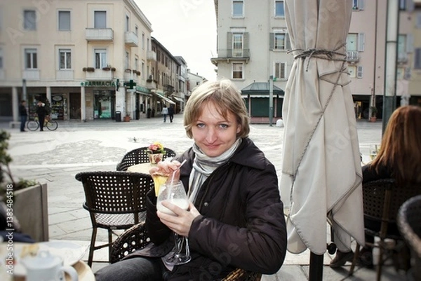 Obraz Woman sitting at a cafe terrace table outdoors and drinking cocktail. Female tourist sitting in a cafeteria taking an aperitif. Smiling woman drinking iced drink in sidewalk cafe. Travel concept