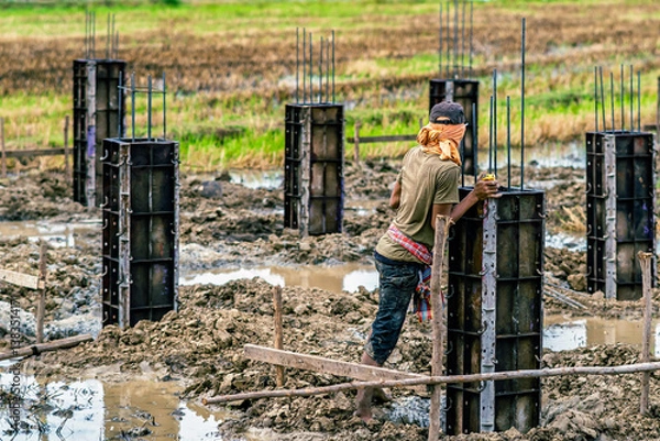 Fototapeta Male worker with Construction of concrete pillars in paddy field. Formwork with reinforcement bar for pad footing. Construction of pad footing foundation. Metal formwork for concrete foundations.