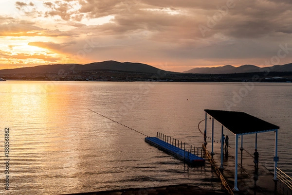 Obraz A dock is on the water with a person standing on it. The water is calm and the sky is orange