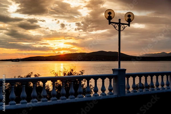 Obraz A beautiful sunset over a body of water with a white fence in the background. The sun is setting and the sky is filled with clouds