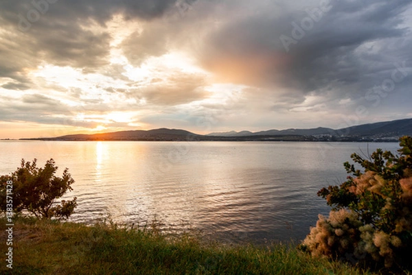 Obraz A beautiful sunset over a calm lake with a few trees in the background. The sky is filled with clouds, creating a moody atmosphere