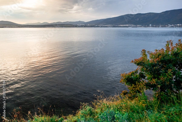 Obraz A calm lake with a tree on the shore. The water is a deep blue color. The sky is cloudy and the sun is setting