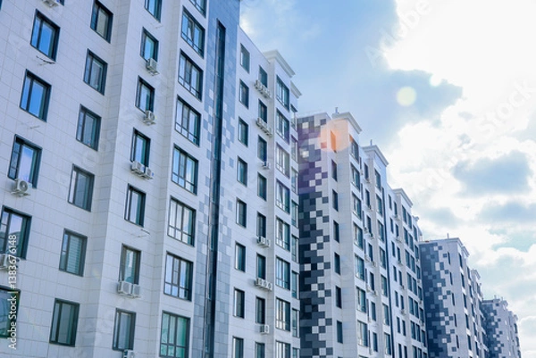Fototapeta The buildings have a modern design with white and gray facades and many windows. Air conditioning is installed on each window. The sky is blue with small clouds. A glare of light from the sun is