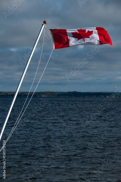 Fototapeta Canadian flag waving on a pole near a serene water body with overcast clouds featuring a natural, serene landscape and scenic surroundings.