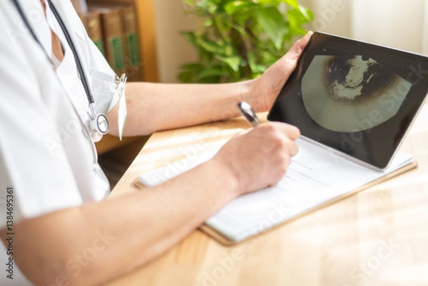 Fototapeta Female doctor in white coat with stethoscope is writing notes on clipboard while examining ultrasound scan displayed on tablet computer, focusing on healthcare and medical diagnosis