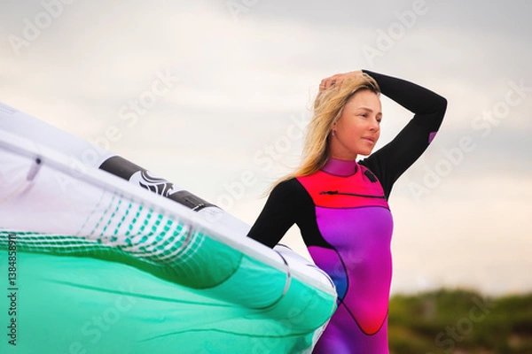 Obraz surfer in a wetsuit ponders the weather on a sandy beach while standing in the sand inflating a kite. A sporty girl prepares her equipment before riding