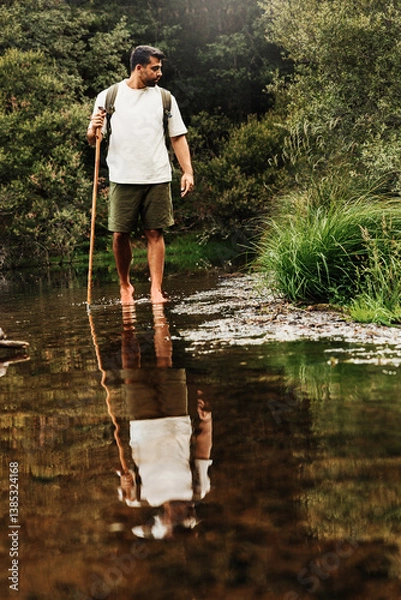 Fototapeta Hiker crossing a stream barefoot in a green forest with a walking stick and backpack