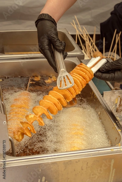 Obraz Chef holds tornado potato or spiral chips in boiling oil in fryer with tongs. Street food. Close-up