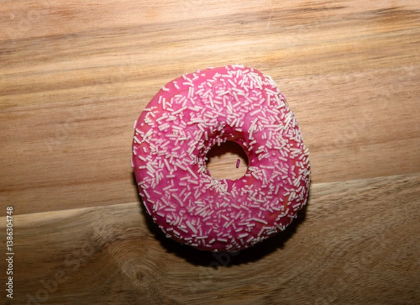 Obraz A vibrant pink frosted donut adorned with white sprinkles rests on a light wooden surface, creating a tempting treat perfect for a sweet snack any time of day
