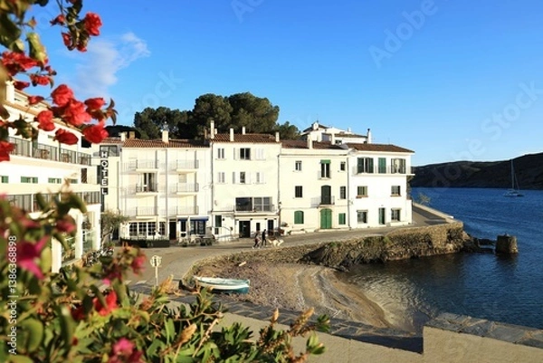 Fototapeta View of Cadaqués, a Mediterranean seaside town in springtime featuring small wooden boat on Es Pianc beach with people walking along promenade against backdrop of whitewashed buildings