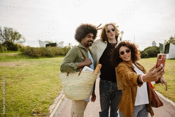 Obraz Three multiethnic friends in their late 20s are standing in a park and taking a selfie using a smartphone held by the Latina woman in a brown jacket, with the men standing behind her, on a sunny day.