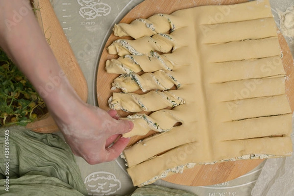 Obraz Top view woman's hand twists dough strips on wooden board. Green towel, microgreens sprouts on kitchen table. Home cooking baking puffs