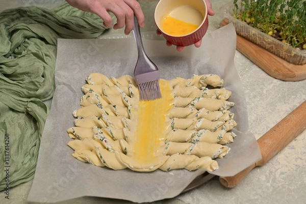 Obraz Woman's hands smears yolk on the twisted dough strips with a brush. Red small bowl with egg. Rolling pin, green towel, microgreens sprouts on kitchen table. Home cooking baking puffs