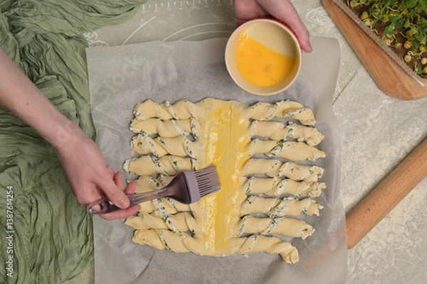Obraz Top view woman's hands smears yolk on the twisted dough strips with a brush. Red small bowl with egg. Rolling pin, green towel, microgreens sprouts on kitchen table. Home cooking baking puffs