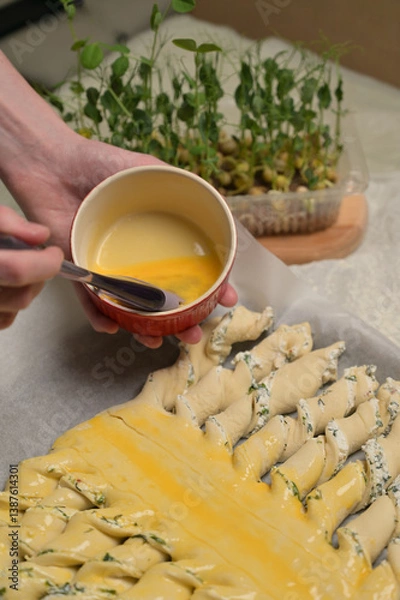 Obraz Close up woman's hands smears yolk on the twisted dough strips with a brush. Red small bowl with egg. Rolling pin, microgreens sprouts on kitchen table. Home cooking baking puffs