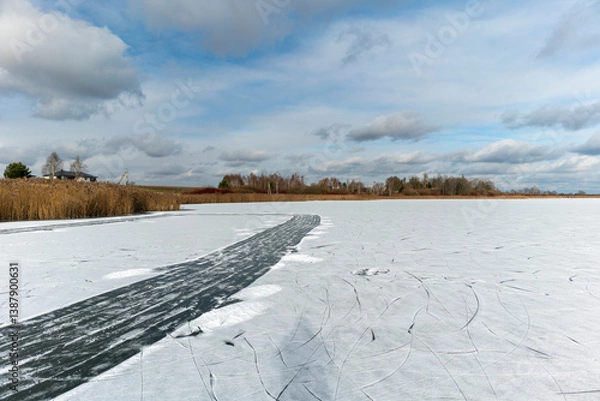 Fototapeta Frozen Lake with Cracked Ice and Winter Landscape. Scenic winter landscape featuring a frozen lake with a prominent crack or ice path cutting through the snow-covered surface. In the distance, dry ree