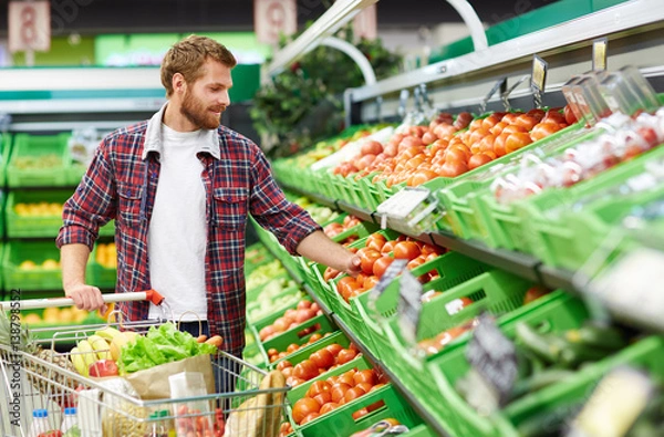 Obraz Przystojny mężczyzna z wózkiem na zakupy dotykający pomidora w celu określenia jego jakości w dziale owoców i warzyw w supermarkecie