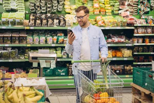 Fototapeta Smiling customer using phone and pushing shopping cart in supermarket