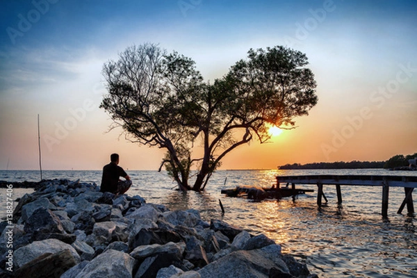 Fototapeta man in frustrated depression sitting alone on the rock dam extended into the sea and looking at tree in the shape of heart on sunset., the concept of lonely, sadness, depressed and broken heart.