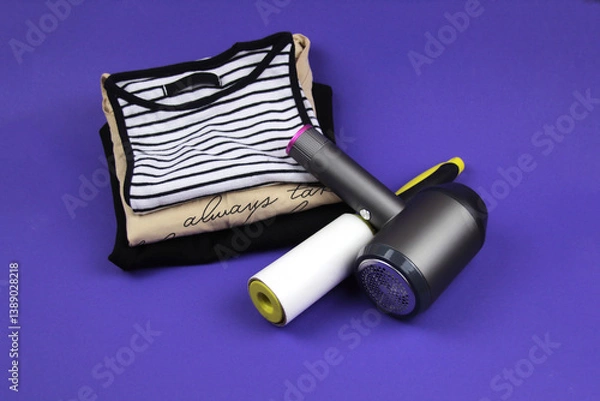 Fototapeta Clothes roller and lint machine are lying on clothes and purple background. In the background are several T-shirts of different colors.