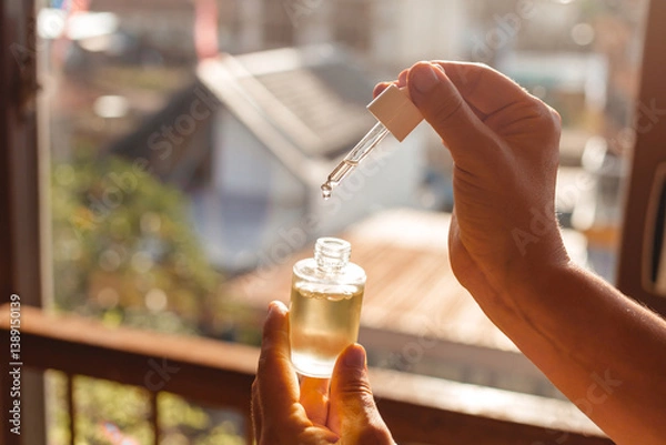 Fototapeta Female hands holding a glass bottle of hair and skin care oil against the sun's rays. Glass pipette with a drop of moisturizing face serum with oils