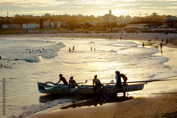 Obraz Team of girls push a surfboat in the water at bondi beach sourranding by the sunset light