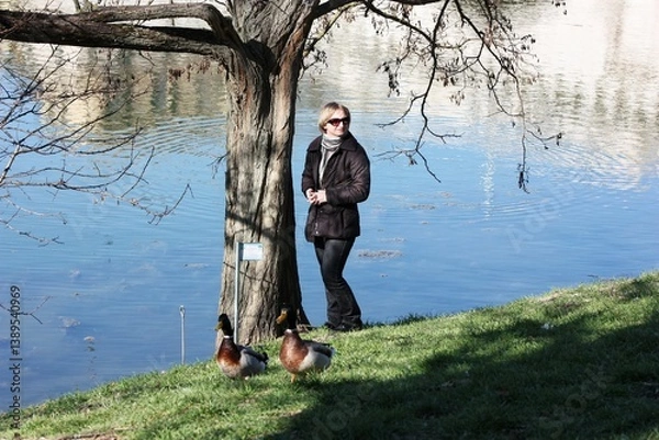 Obraz Young woman in sunglasses walking by the duck pond. Young girl walks by a lake in the autumn park. Adult female with short hair walks in on spring park by a river. Young woman resting near lake