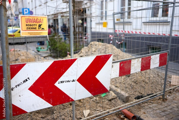 Obraz A fenced construction zone in a European city street, marked by directional warning signs and excavation work. The sign in Polish alerts pedestrians to ongoing ground works.