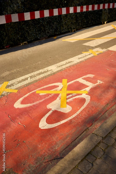 Obraz A red bike lane marked with yellow X tape, signaling closure or restriction. Urban textures, cracked asphalt, and morning light tell a silent story of change and interruption.