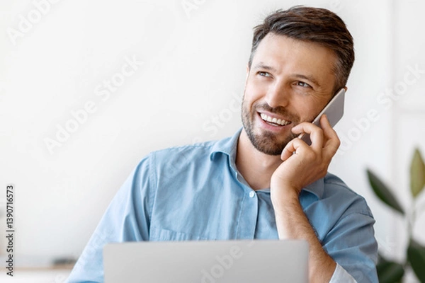 Fototapeta Handsome Male Entrepreneur Having Phone Conversation And Drinking Coffee At Workplace, Sitting At Desk With Laptop In Office, Having Break At Work, Enjoying Mobile Communication, Copy Space