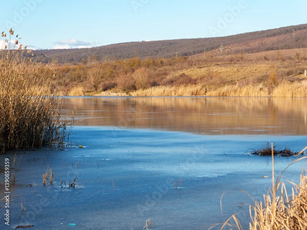 Fototapeta A serene river landscape features a thin layer of ice covering the water. Tall golden reeds line the riverbank under a bright blue sky, with distant hills adding depth to the scene.