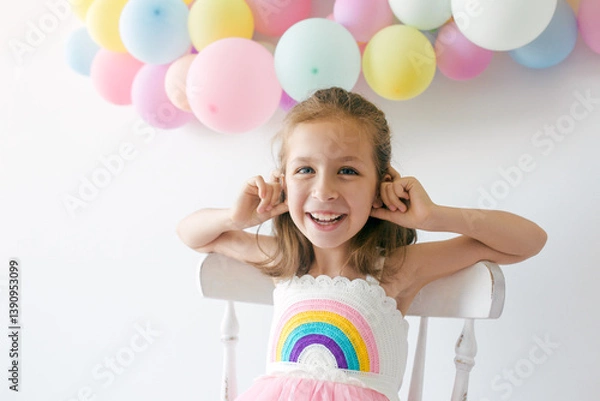 Fototapeta Portrait of little happy girl in dress, sitting on the chair with colorful balloons on a white background, studio shoot. Celebrations, birthday and playing concept.