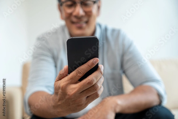 Obraz Handsome asian mature old man using smartphone on sofa in living room at home. Happy Portrait of cheerful smiling senior asian man holding cell phone. Mature People and lifestyle
