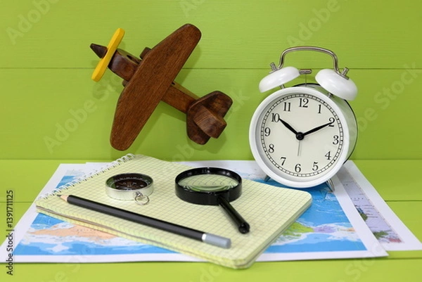 Fototapeta A still life about time to travel on the green wooden background  with retro alarm clock, wooden retro model of airplane, world maps, compass, magnifier, notebook and pencil. Concept of travel.