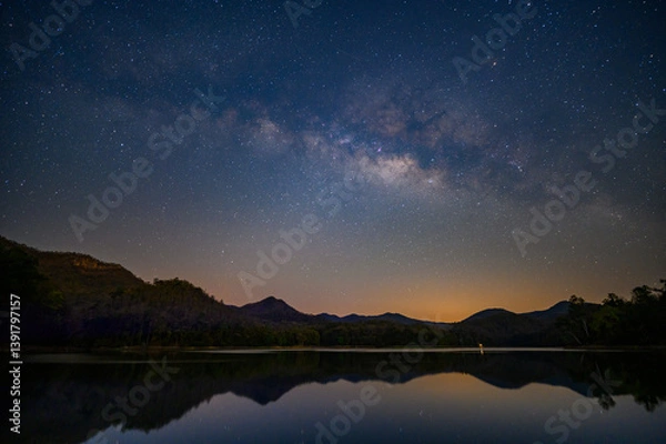 Fototapeta milky way stretches across the star-filled night sky above dark mountain silhouettes and a calm reflective lake, with a soft orange glow near the horizon