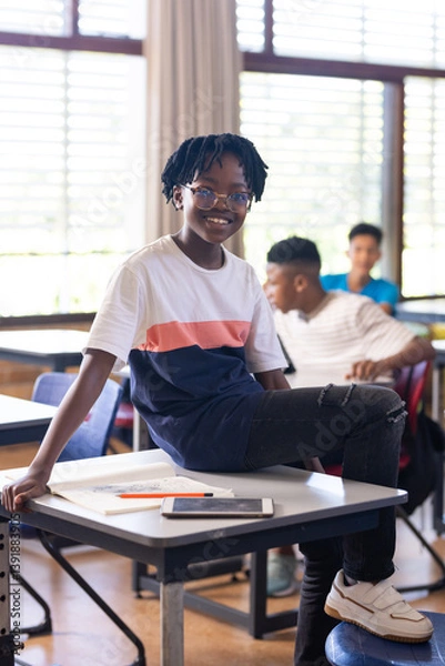 Obraz In school, smiling student sitting on desk with tablet and notebook nearby