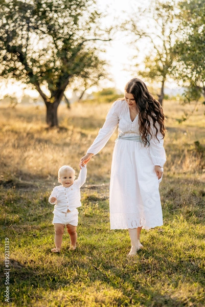 Fototapeta Mother and son in white clothes walking and playing in the garden in summer. Family time, childcare and parental love in nature