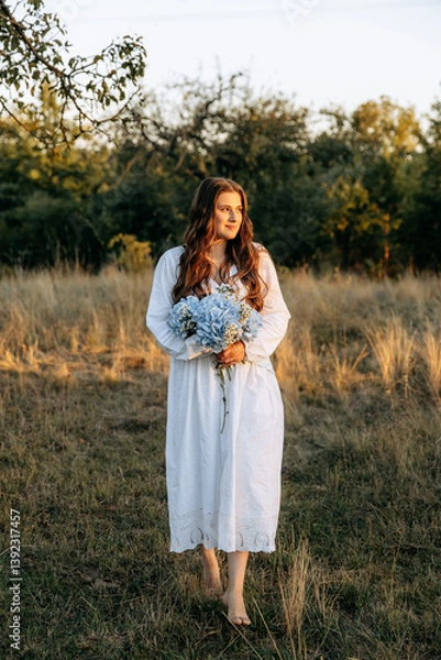 Obraz Portrait of a cute girl in a white dress barefoot in a garden with blue hydrangea. Summer walks in nature under sunlight
