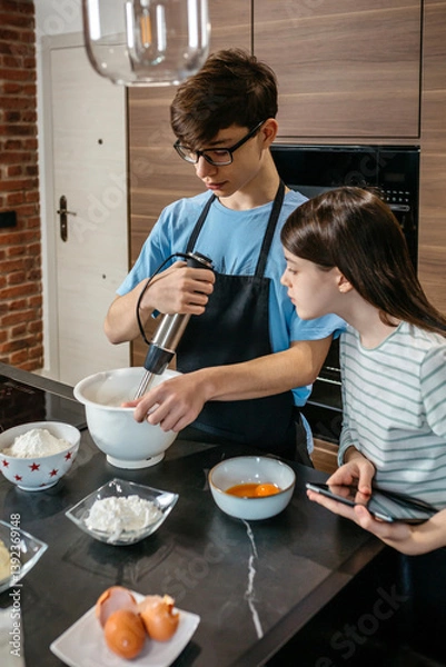 Fototapeta Teenage brother mixing ingredients for a cake using hand blender while curious sister looking inside of the bowl and holding tablet with recipe in kitchen. Siblings learning culinary skills concept.