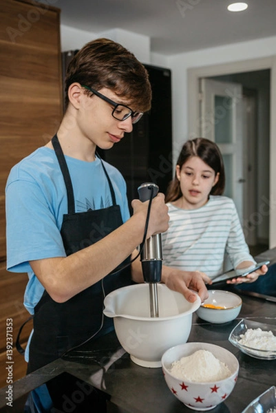 Fototapeta Teenage boy using hand blender while mixing ingredients in bowl, following recipe on tablet held by sister in kitchen, enjoying family moment and learning culinary skills. Happy time cooking concept.